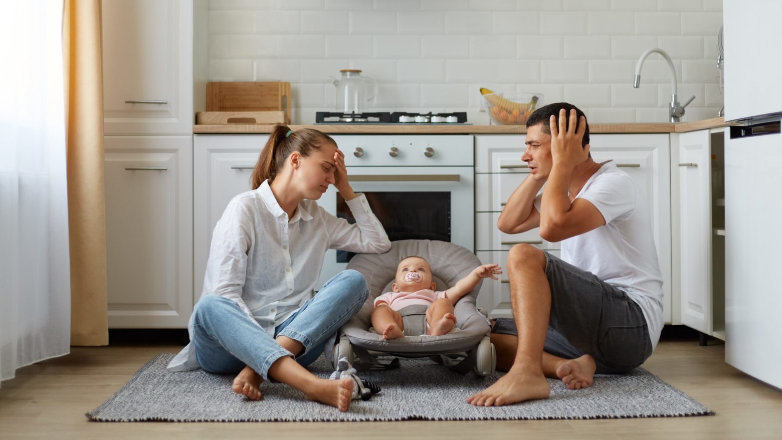 Portrait of mother and father sitting on floor in kitchen with little son or daughter in rocking chair on kitchen floor, tired sleepless parents keeping hands on head, need rest. Starševstvo <em>presega</em> naše <strong>moči</strong>